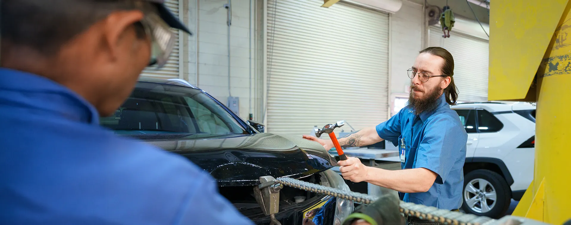 Student uses a slide hammer to pull a dent from a car panel while another person observes in an auto repair shop
