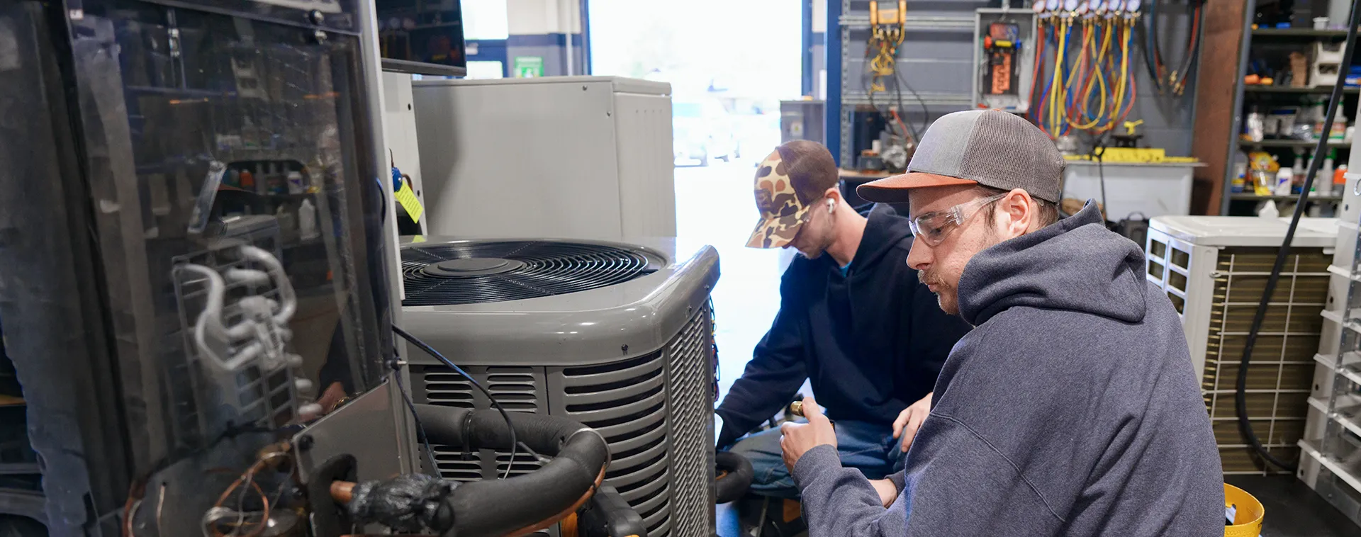 Two people in a workshop working on a large HVAC unit, with tools, wiring, and equipment visible around them.