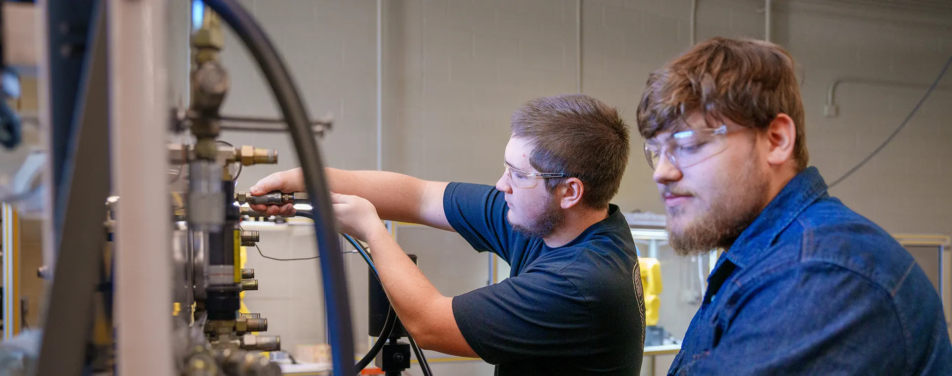 Two people wearing safety glasses adjust valves and gauges on industrial equipment with hoses and metal piping.