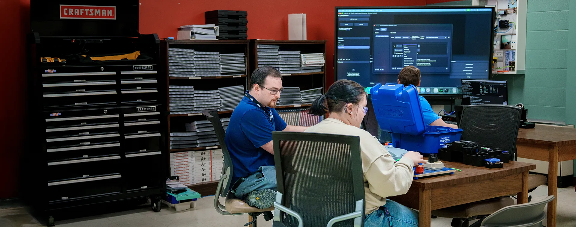 Students seated at desks work with tools and devices while a large screen displays software in a classroom with red walls and tool cabinets.