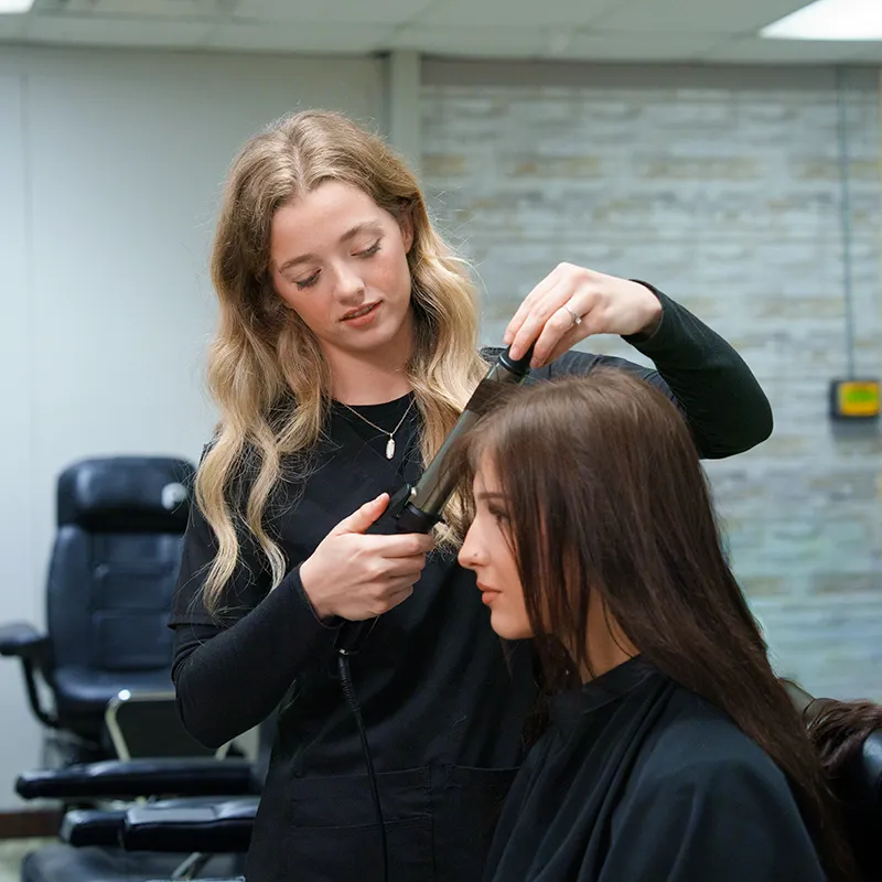 Student uses a curling iron to style another person&rsquo;s hair in a salon classroom with chairs and mirrors in the background.