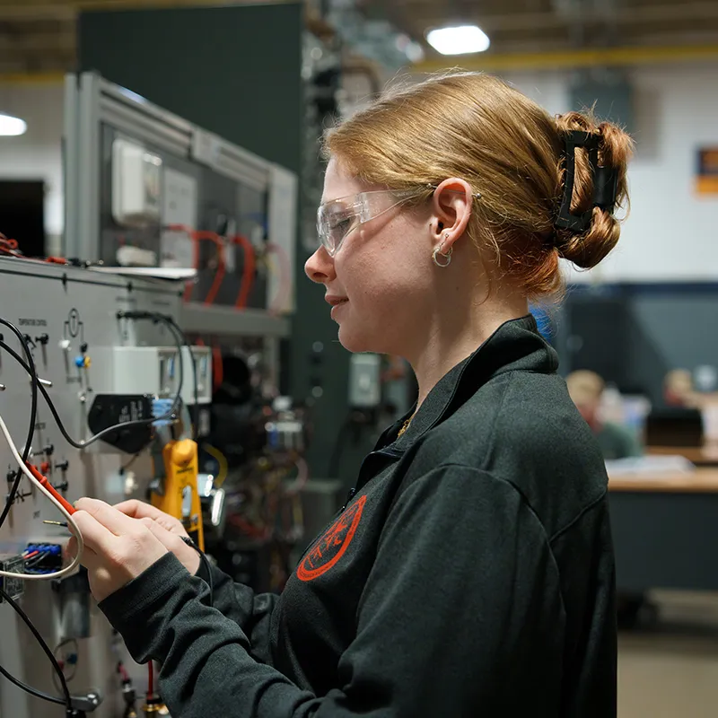 Person wearing safety glasses connects wires on an electrical training panel with meters, switches, and cables in a lab setting.