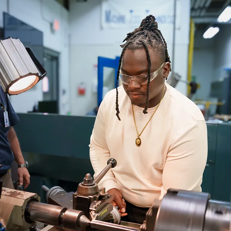 Student wearing safety glasses operates a metal lathe, adjusting controls under a task light in a workshop.