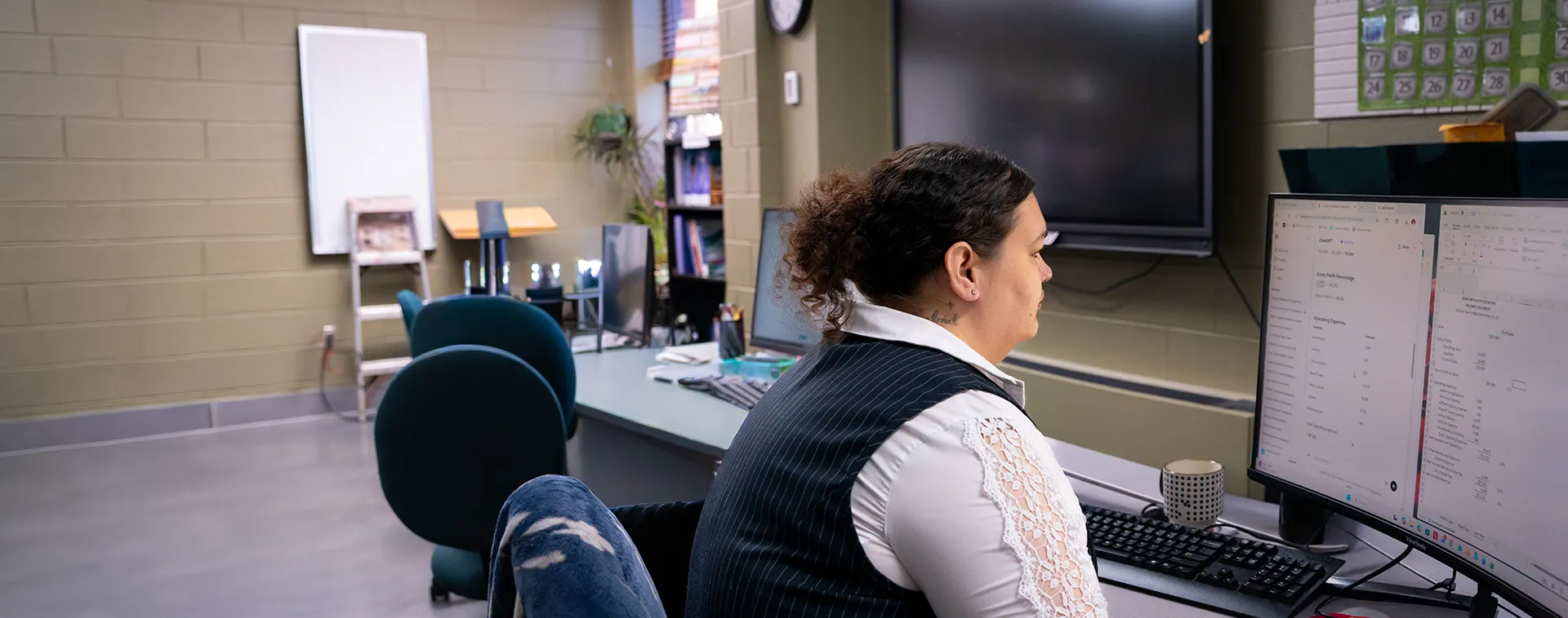 Person seated at desk using dual monitors in a classroom office; whiteboard, shelves, and chairs in background.