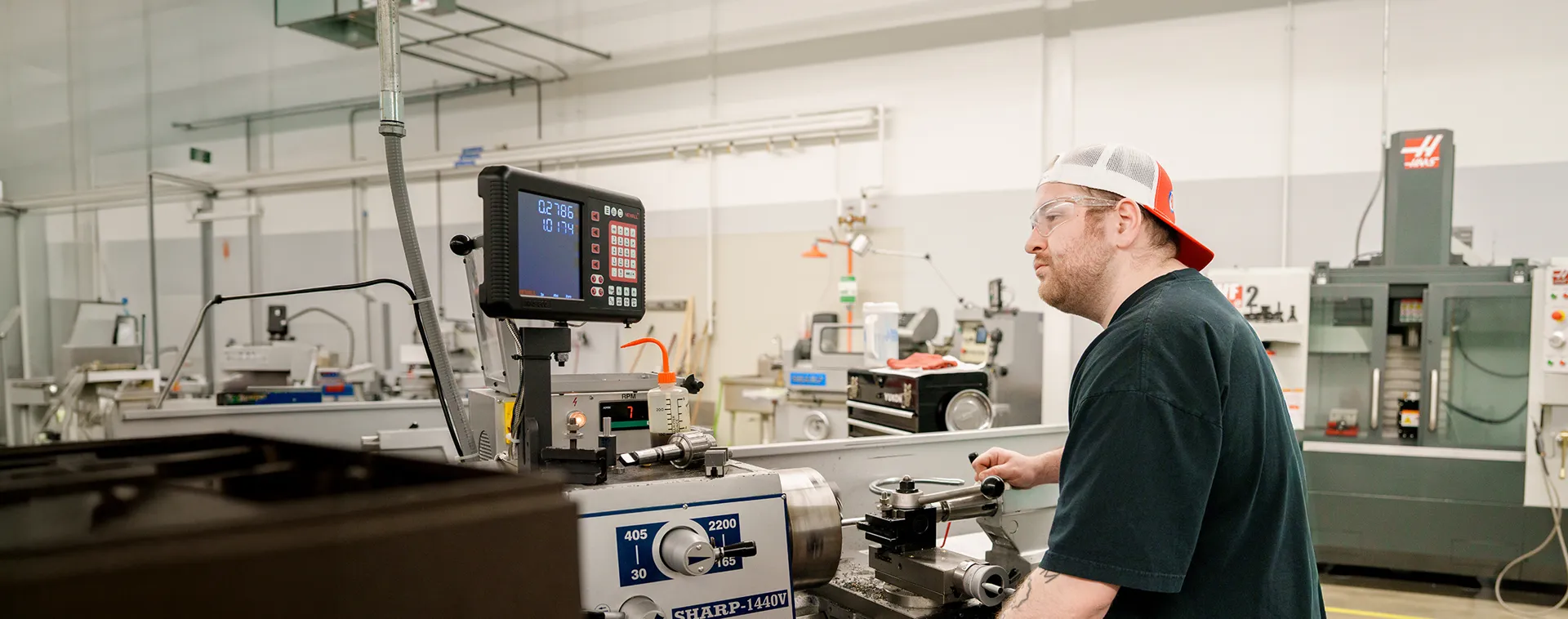 Student uses a lathe machine with digital display in a large shop filled with industrial equipment.