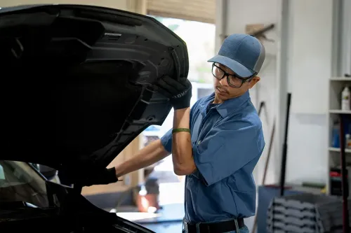 Technician wearing gloves lifts a car hood inside a repair bay with tools and equipment in the background