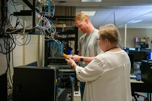 Two people stand beside a server rack with cables, one holding a tool while working on network wiring in a computer lab.