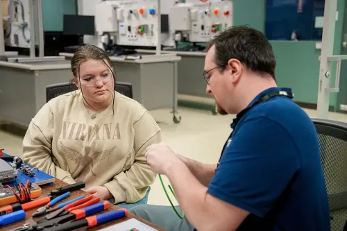 Instructor demonstrates wire stripping to a student at a table with hand tools and cables, with lab equipment in the background.
