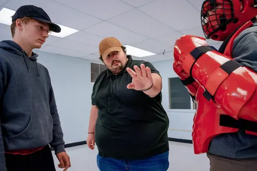 Instructor gestures while two students stand nearby, one wearing red padded protective equipment during a training exercise.